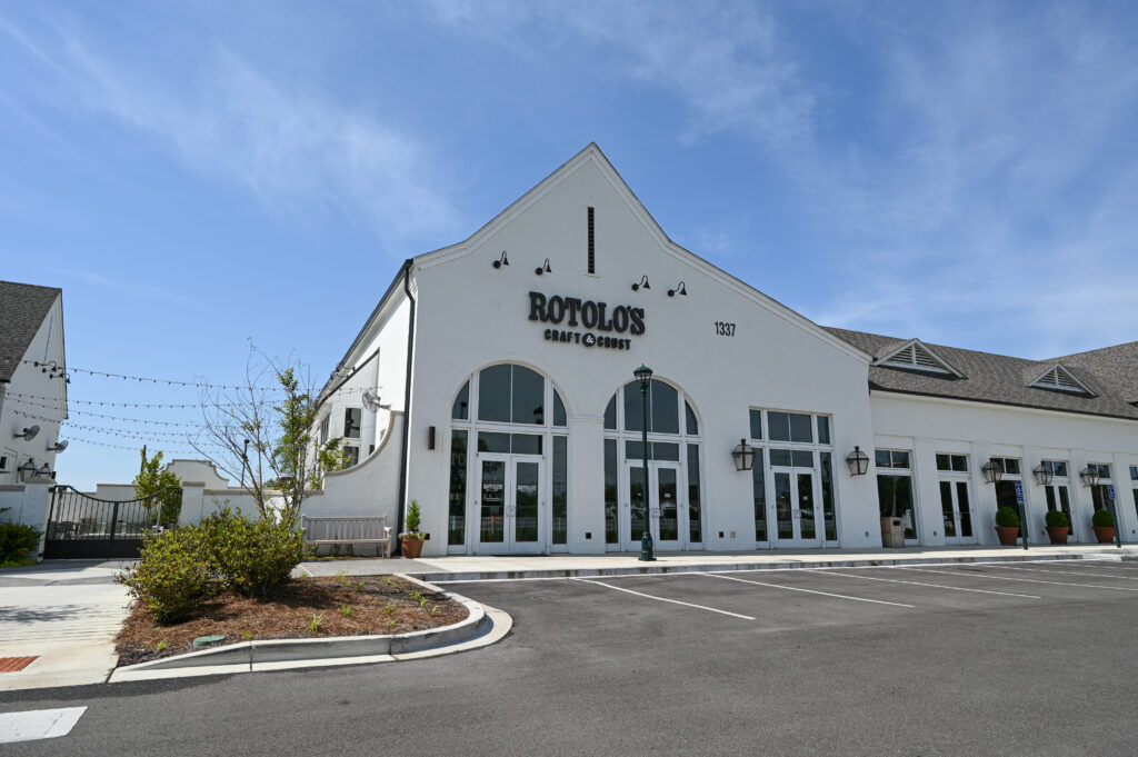 A white building with arched windows and "Rotolo's Craft Corner" sign, home to handcrafted pizza and Detroit-style pizza, with an empty lot in front.