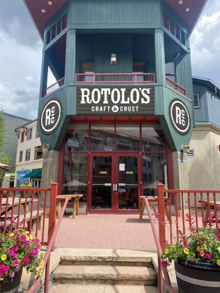 Front entrance of Rotolo’s Pizzeria with large sign above glass doors, flower planters, red railings, and a hint of handcrafted pizza inside.