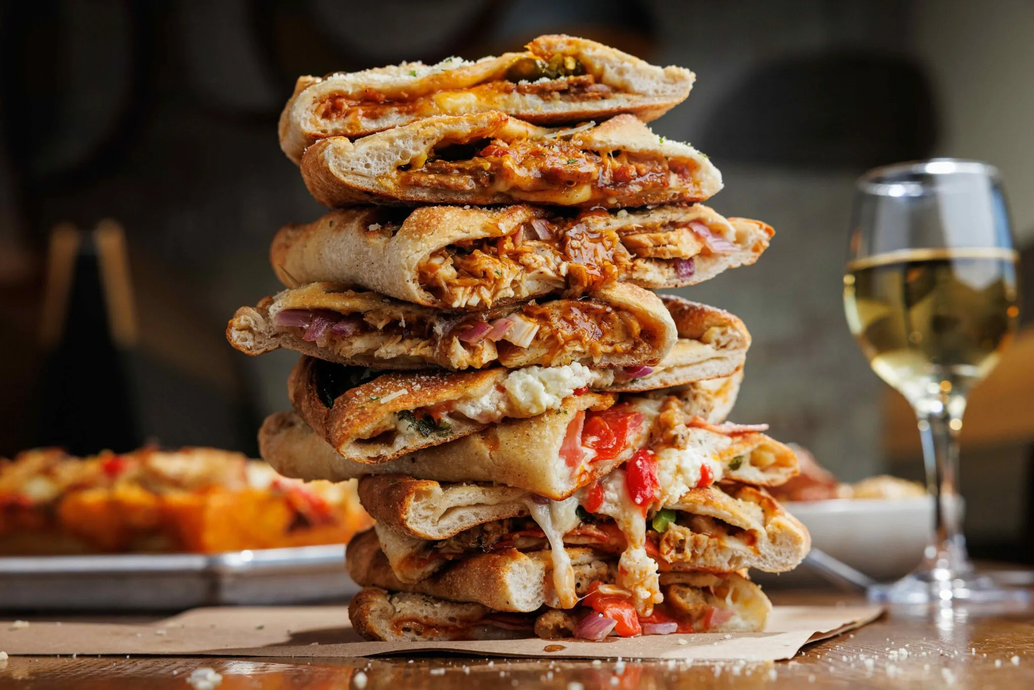A tall stack of assorted stuffed calzones from Rotolo’s, celebrating 30 years of tradition, with wine on a wooden table.